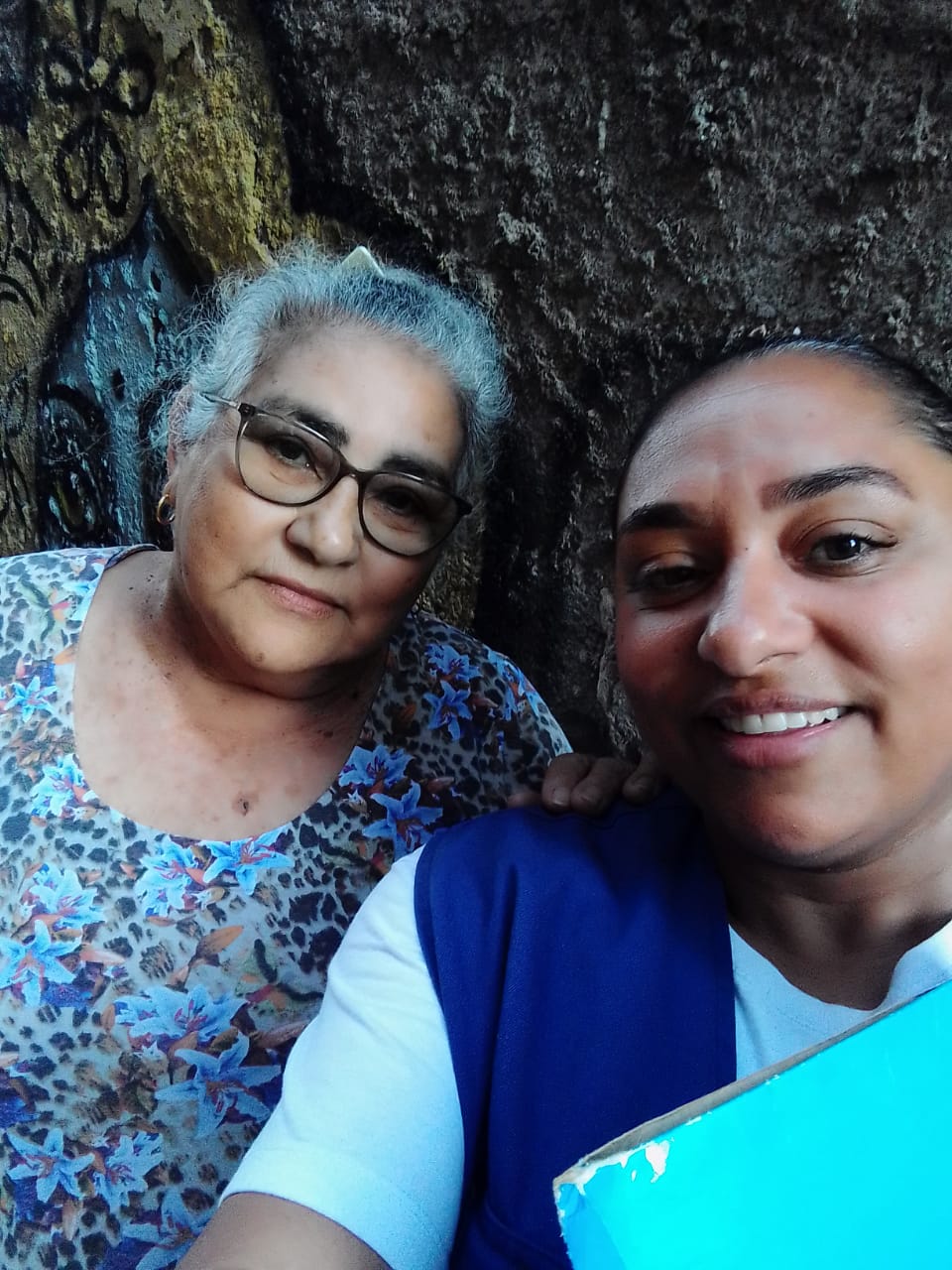 A foto mostra Josélia, uma mulher jovem de pele parda, usando um colete azul; ao seu lado e com a mão em seu ombro está uma moradora visitada pela agente de saúde. É uma mulher de cabelos brancos, que usa óculos e uma blusa estampada em tons de azul